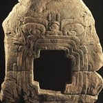 Large olmec stone carving against a black background. It is in the shape of an earth monster with a cross shaped mouth. It is the piece described in the main body text.