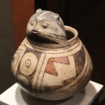 An ancient ceramic vessel. White background with geometric black and red designs. It has a ceramic animal head projecting from the rim.